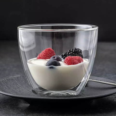 Un vaso de vidrio de doble pared está lleno de yogur cremoso y adornado con frambuesas, moras y arándanos frescos. El vaso está sobre un plato negro con una cuchara al lado, delante de un fondo oscuro.