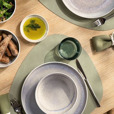 Vista superior de una mesa de comedor con platos grises, manteles individuales verdes, un cuenco de palitos de pan, un cuenco de salsa verde de eneldo y un pequeño vaso de color verde. Las servilletas están enrolladas cuidadosamente y colocadas junto a los platos.