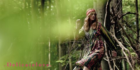 Una mujer con un vestido de estampado colorido y una cinta roja para el cabello está de pie en medio de un exuberante bosque verde. En la esquina inferior izquierda, las palabras "Delicatelove" están escritas en rosa. El primer plano está borroso con hojas verdes.