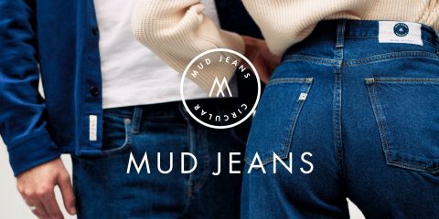 Dos personas están de pie muy cerca una de la otra, vistiendo vaqueros azules de Mud Jeans. La persona de la izquierda lleva una chaqueta y la de la derecha muestra la parte trasera de los vaqueros. El logotipo y el texto de Mud Jeans son claramente visibles.