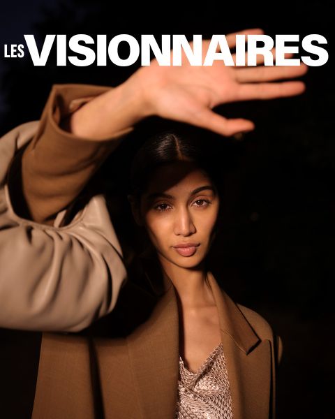 Una persona con un abrigo marrón levanta una mano para cubrir parcialmente su rostro. Está de pie frente a un fondo oscuro. En la parte superior de la imagen, las palabras "Les Visionnaires" están escritas en letras blancas y negritas.