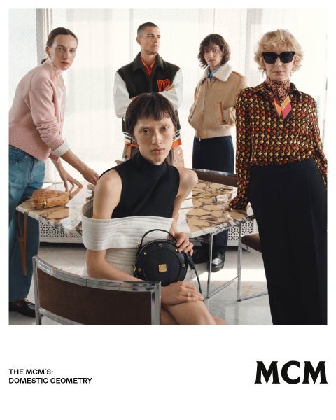 Cinco personas elegantes posan en un interior alrededor de una mesa de mármol, presentando bolsos y moda de MCM en un ambiente moderno y elegante.