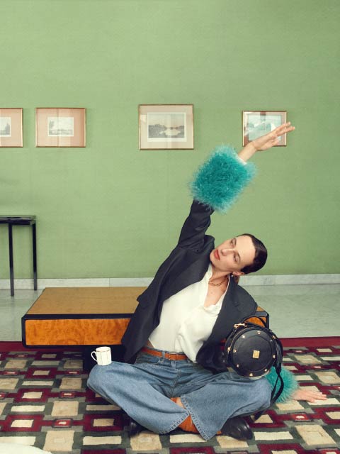 Una persona en jeans y un blazer con puños azules y peludos posa con el brazo levantado en una habitación elegante con paredes verdes, obras de arte enmarcadas y una alfombra estampada.