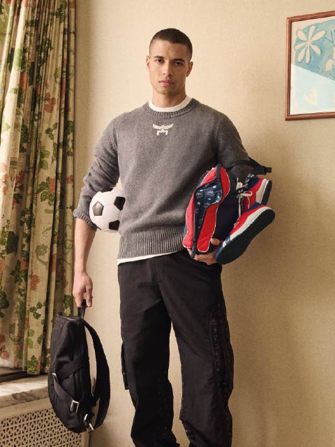 Un joven con un suéter gris sostiene una mochila negra y zapatillas de deporte en la mano, un balón de fútbol bajo el brazo, y está de pie en el interior cerca de unas cortinas floreadas.