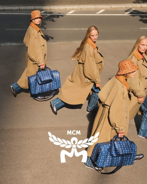 Tres mujeres con abrigos de color marrón claro y sombreros naranjas cruzan una calle, llevando bolsos azules de MCM con un logo blanco de MCM en primer plano. La imagen tiene un efecto espejo que hace que la escena parezca duplicada.