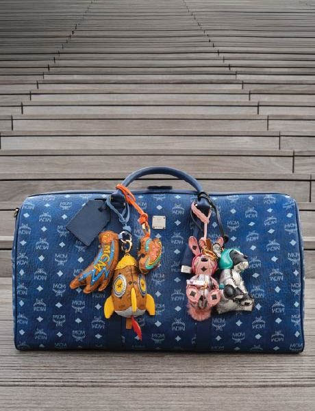Un bolso de diseño MCM azul con un patrón de logotipo blanco está sobre escalones de madera adornados con coloridos llaveros de animales y objetos, incluyendo un cohete, un conejo y un oso.