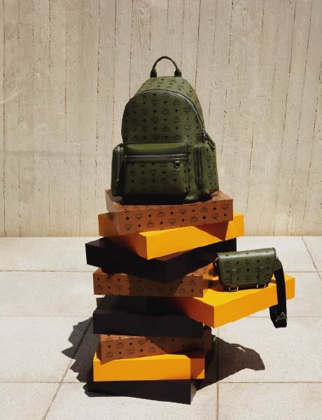 Una mochila de diseño verde y un bolso bandolera a juego están sobre una pila de cajas naranjas y marrones frente a una pared con textura beige y un suelo de baldosas.