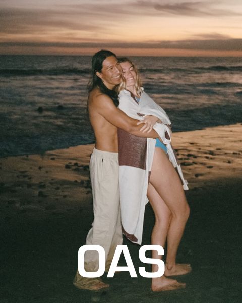 Una pareja se abraza al atardecer en una playa. El hombre está sin camisa y lleva pantalones ligeros, mientras que la mujer está envuelta en un chal blanco y marrón. En el fondo se ven las olas del mar, debajo está el texto "OAS".