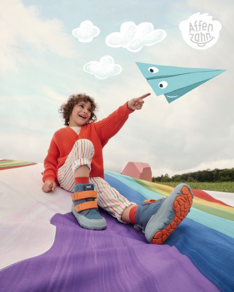 Un niño con un suéter naranja brillante y pantalones a rayas está sentado en una superficie colorida y señala un avión de papel estilo cómic en el cielo. Sonríe y está rodeado de nubes esponjosas. En la esquina pone "Affenzahn".