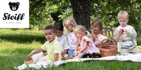 Un grupo de niños sentados en una manta al aire libre, rodeados de árboles. Están jugando con cestas y animales de peluche. El logo de Steiff está en la esquina superior izquierda.