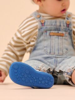 Un niño pequeño con camisa a rayas y mono de mezclilla está sentado con una pierna al aire libre, mostrando la suela azul texturizada de su zapato.