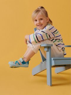 Niño sonriente con camisa a rayas y pantalones claros sentado de lado en una silla de madera azul sobre un fondo amarillo.