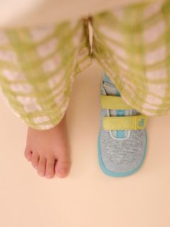Un pie desnudo de niño junto a una zapatilla deportiva con correas amarillas, ambos visibles debajo de un pantalón de cuadros verde claro sobre un suelo beige.