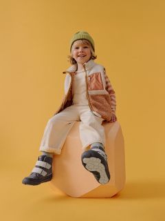 Un niño sonriente con un gorro verde, camisa a rayas y chaleco está sentado sobre un bloque de color melocotón frente a un fondo amarillo, vistiendo botas y pantalones claros.