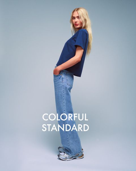 Una persona con cabello largo y rubio está de pie de lado frente a un fondo azul. Lleva una camiseta azul oscuro, vaqueros azul claro y zapatillas de deporte. El texto "COLORFUL STANDARD" se muestra en blanco.