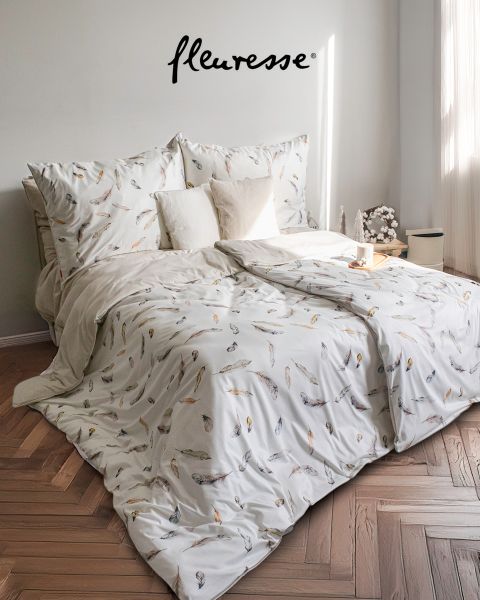 Un dormitorio acogedor con una cama bien hecha con ropa de cama blanca con un diseño de pájaros. Dos almohadas decorativas están apoyadas en el cabecero. La luz del sol entra a través de cortinas transparentes, proyectando un brillo suave sobre el suelo de madera clara.