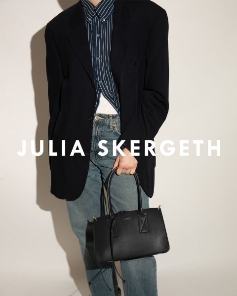 Persona con un blazer negro sobre una camisa a rayas y jeans azules, sosteniendo un bolso negro. El texto "JULIA SKERGETH" es claramente visible superpuesto a la imagen.