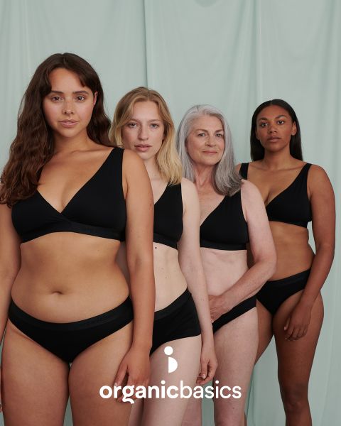 Cuatro mujeres de diferentes edades y figuras están de pie con confianza frente a un fondo verde claro, en ropa interior negra a juego. En el borde inferior de la imagen está el texto "organicbasics".