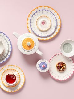Platos coloridos con estampado floral, una tetera, una taza con té y limón, y una pieza de repostería, dispuestos sobre un fondo rosa.