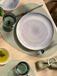 Puesta de mesa con un mantel individual verde, un plato de cerámica, un vaso, un tenedor, un cuchillo, un pequeño cuenco con aceite y eneldo, y una servilleta de tela verde con un servilletero.