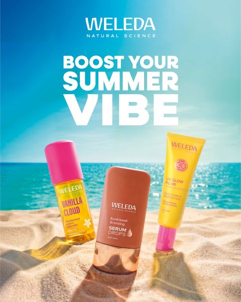 Tres productos de cuidado de la piel Weleda en la arena con el mar y el cielo azul de fondo. Texto: "Boost Your Summer Vibe". Escena de playa luminosa y veraniega.