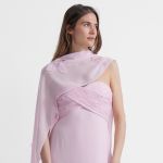 Eine Frau in einem blassrosa, trägerlosen Kleid mit einem über eine Schulter drapierten transparenten Schal steht vor einem weißen Hintergrund und blickt zur Seite.