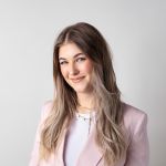 Mujer con cabello castaño claro largo sonríe suavemente y lleva un blazer rosa claro sobre una blusa blanca frente a un fondo blanco sencillo.