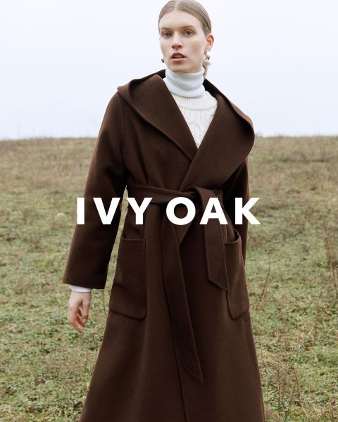 Una persona con un abrigo largo marrón con cinturón está de pie en un campo abierto. Debajo lleva un jersey de cuello alto blanco y mira hacia adelante. El texto "IVY OAK" está superpuesto a la imagen. El fondo es un paisaje herboso y nublado.