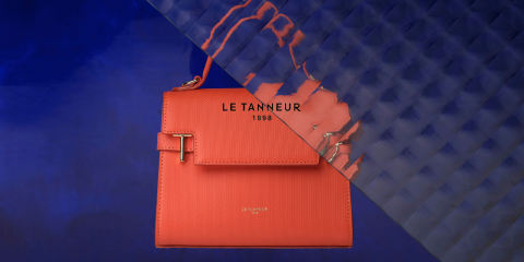 Un elegante bolso naranja de Le Tanneur se encuentra frente a un fondo texturizado de color azul brillante. El bolso tiene un diseño elegante con cierre de solapa y el nombre de la marca "Le Tanneur 1898" grabado.