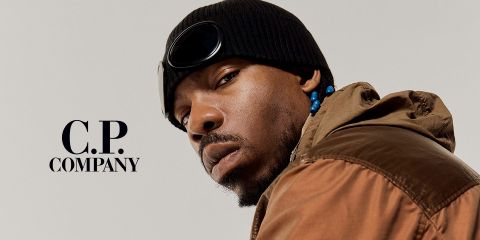 Hombre con chaqueta marrón, gorro negro con gafas protectoras y cuentas en el pelo. Mira a la cámara con expresión seria. En el lado izquierdo, sobre un fondo sencillo, aparece el texto "C.P. Company".
