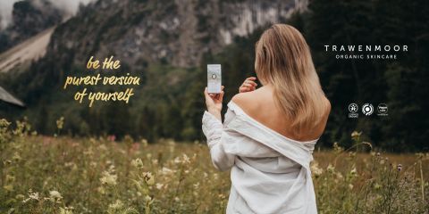 Una mujer con cabello largo y rubio está de pie en un campo con la cara girada y sostiene un producto para el cuidado de la piel en la mano. Lleva un top con los hombros descubiertos. El texto dice "Sé la versión más pura de ti mismo". En el fondo se ven montañas y hierba. En la pantalla se lee "TRAWENMOOR CUIDADO ORGÁNICO DE LA PIEL".