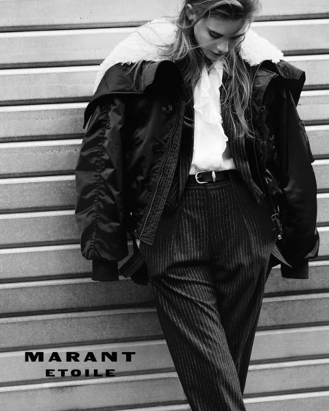 Fotografía en blanco y negro de una mujer con una chaqueta con cuello de lana de oveja, una blusa con volantes y pantalones de raya diplomática, apoyada contra una pared de metal. El texto "MARANT ETOILE" está en la parte inferior izquierda.