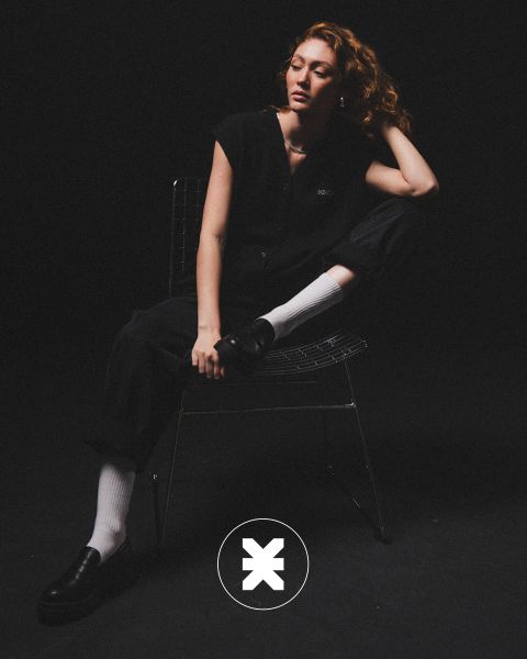 Una persona con cabello rizado está sentada en una silla de alambre en una habitación oscura, vestida con ropa negra, calcetines blancos y zapatos negros. Una pierna está doblada, un codo descansa sobre la rodilla y en el suelo hay un logo circular con una "X".