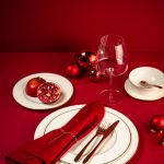 Eine festliche Tischdekoration mit rotem Thema, bestehend aus einer roten Serviette, goldenem Besteck, einem weißen Teller und einem Granatapfel. Ein Weinglas und rote Ornamente werten das Dekor auf. Der Hintergrund und der Tisch sind in kräftigem Rot gehalten und schaffen eine elegante Feiertagsatmosphäre.
