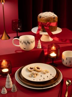 Una mesa festiva con un gran pastel atado con un lazo rojo y espolvoreado con azúcar glas en un soporte. Hay galletas en platos, tazas temáticas, velas, adornos navideños rojos y blancos y un fondo rojo intenso.