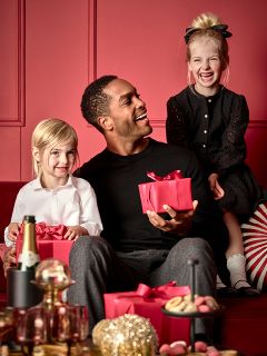 Un hombre está sentado con una caja de regalo roja en un sofá rojo y sonríe a dos niños pequeños. Un niño, vestido de blanco, sostiene un regalo rojo similar, mientras que el otro, vestido de negro, está sentado en el respaldo del sofá. En primer plano hay una mesa con golosinas y una botella.