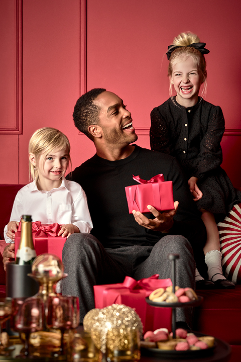 Un hombre está sentado con una caja de regalo roja en un sofá rojo y sonríe a dos niños pequeños. Un niño, vestido de blanco, sostiene un regalo rojo similar, mientras que el otro, vestido de negro, está sentado en el respaldo del sofá. En primer plano hay una mesa con golosinas y una botella.
