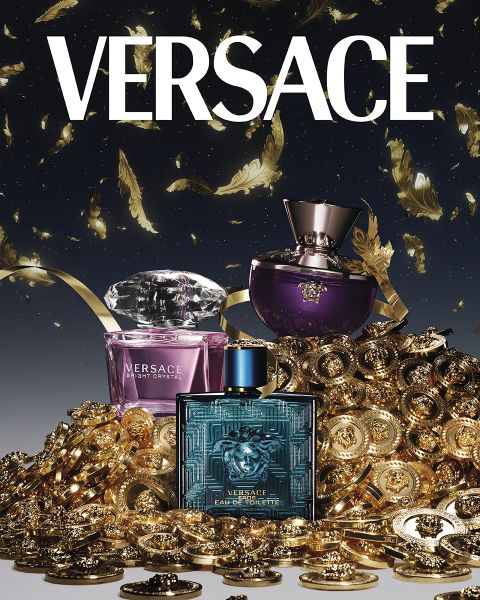 Frascos de perfumes Versace, Bright Crystal, Dylan Purple y Eros Eau de Toilette, rodeados de un montón de monedas de oro y plumas. El logo de Versace se ve claramente en la parte superior sobre un fondo oscuro.