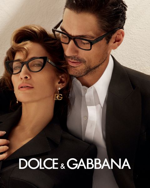 Una mujer y un hombre con gafas elegantes se inclinan cerca el uno del otro. Ella lleva pendientes de DG y un conjunto negro, él lleva un traje negro con camisa blanca. Abajo se ve el logo de Dolce & Gabbana.