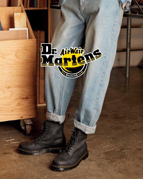 Una persona con vaqueros azul claro y botas con cordones negras Dr. Martens está de pie en el interior sobre un suelo de hormigón. El logotipo "Dr. Martens AirWair" se ve claramente encima de la imagen. En el fondo se ven una estantería de madera y la pata de una silla de metal.