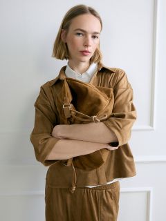 Una mujer con un bob rubio, vestida con un atuendo marrón, está de pie con los brazos cruzados frente a una pared blanca, sosteniendo un gran bolso de gamuza marrón.