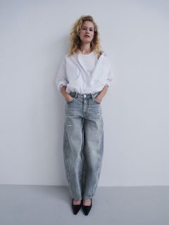 Una mujer con una camisa blanca y jeans grises holgados está de pie con las manos en los bolsillos frente a una pared blanca, lleva zapatos de tacón negros y mira a la cámara.