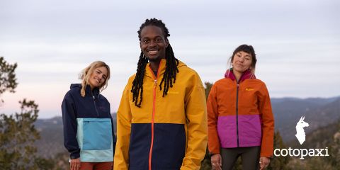 Tres personas con coloridas chaquetas de exterior están de pie en un paisaje natural con árboles y montañas. El cielo está nublado. Abajo a la derecha se ve el logotipo de la marca "Cotopaxi".