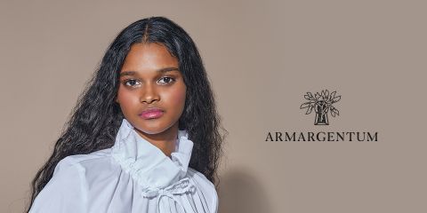 Una mujer con cabello largo y ondulado lleva una blusa blanca con cuello alto. Está de pie frente a un fondo beige, en cuyo lado derecho se encuentra el logo "ARMARGENTUM" con un diseño floral.