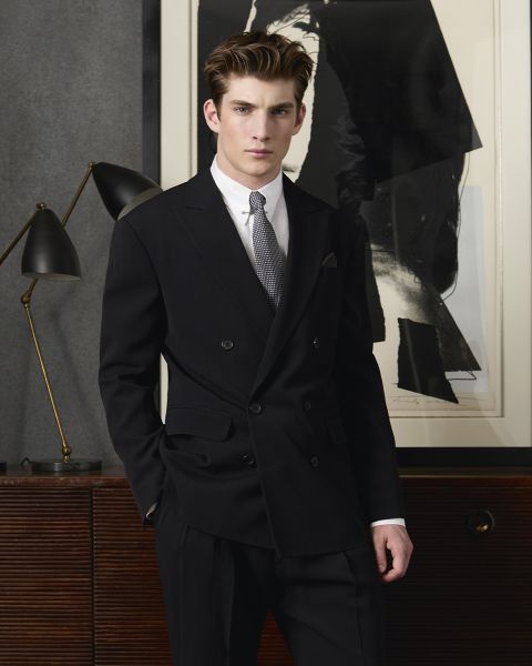 Un hombre con un traje cruzado negro y una corbata gris se encuentra con confianza frente a arte abstracto. A su lado, sobre una superficie de madera, hay una lámpara negra con detalles en latón. El ambiente se ve moderno y elegante.