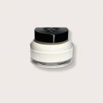 Ein kleines, rundes Glasgefäß mit schwarzem Deckel enthält eine weiße Creme. Der Behälter hat glatte, gewölbte Seiten und steht auf einem schlichten, hellgrauen Hintergrund.