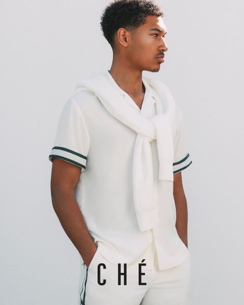 Un hombre con un atuendo blanco con adornos negros, que consiste en una camisa de manga corta y pantalones, posa frente a un fondo blanco liso. Un jersey blanco cuelga sobre sus hombros, con las manos metidas en los bolsillos. Abajo está el texto "CHÉ".