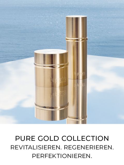 Dos recipientes cosméticos cilíndricos dorados de la "Colección Oro Puro" se colocan sobre una superficie reflectante frente a un tranquilo fondo de agua. El texto dice: "COLECCIÓN ORO PURO. REVITALIZAR. REGENERAR. PERFECCIONAR.".