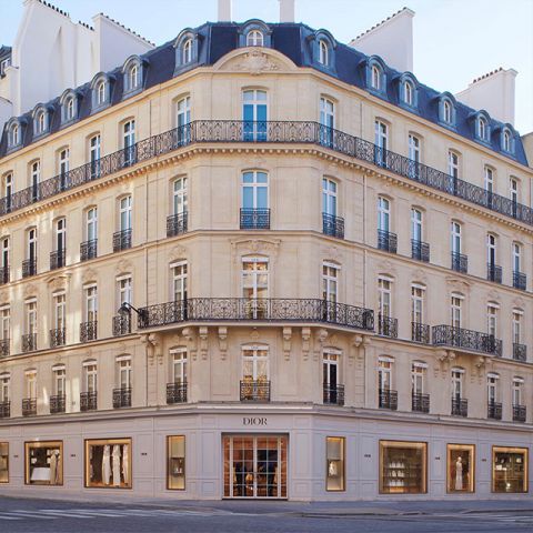 Elegante edificación de esquina con arquitectura clásica, varios pisos, ventanas arqueadas y balcones de hierro forjado ornamentados. En la planta baja se encuentra una tienda Dior con grandes escaparates. La luz natural brillante realza la fachada beige.