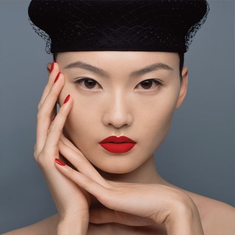Una persona con un llamativo labial y esmalte de uñas rojos posa frente a un fondo gris. Lleva un sombrero negro con textura de red y sus manos están elegantemente colocadas alrededor de su rostro, acentuando el maquillaje brillante.